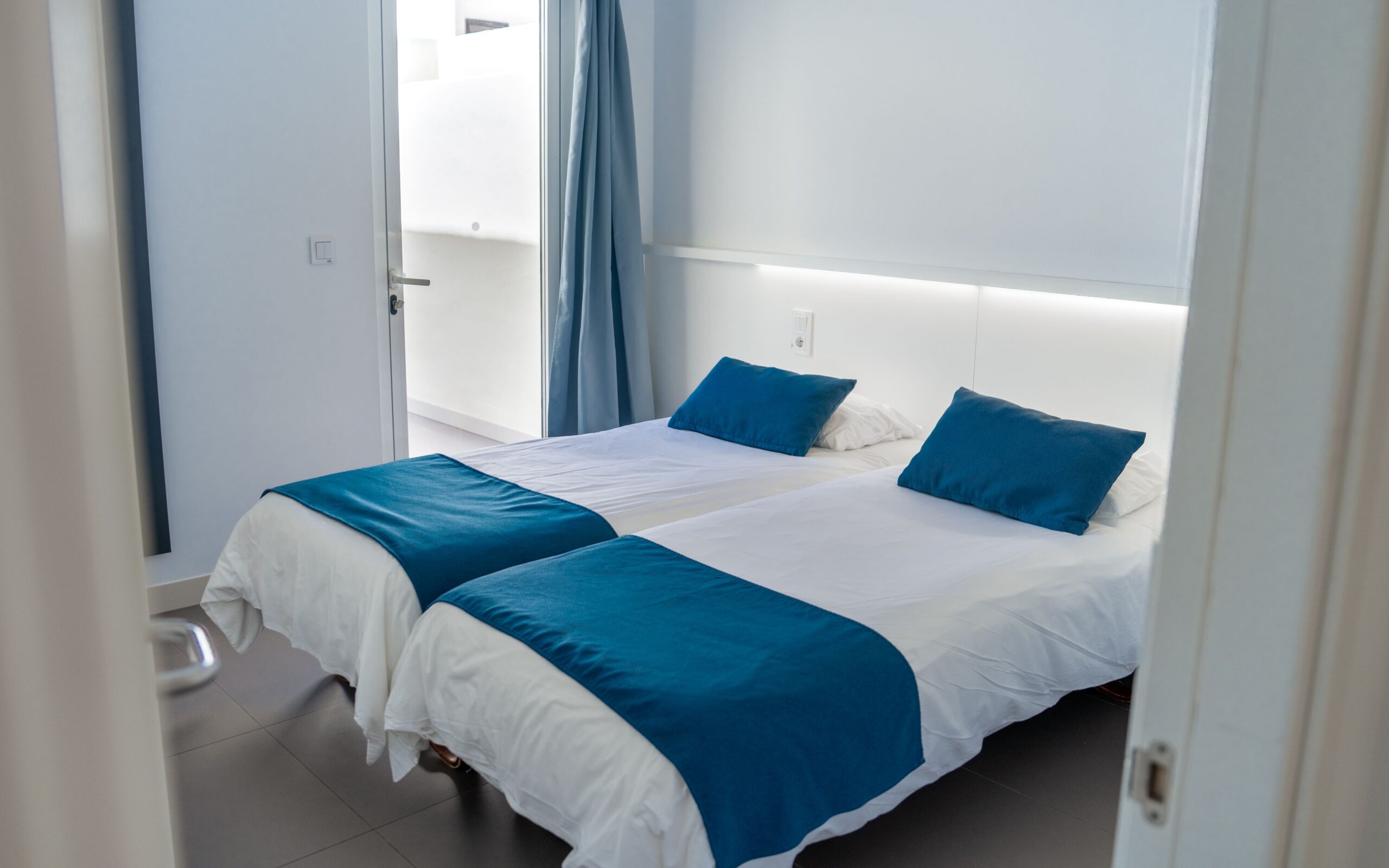 2 single beds with white and blue beddng in a bedroom with white walls and a glass patio door.