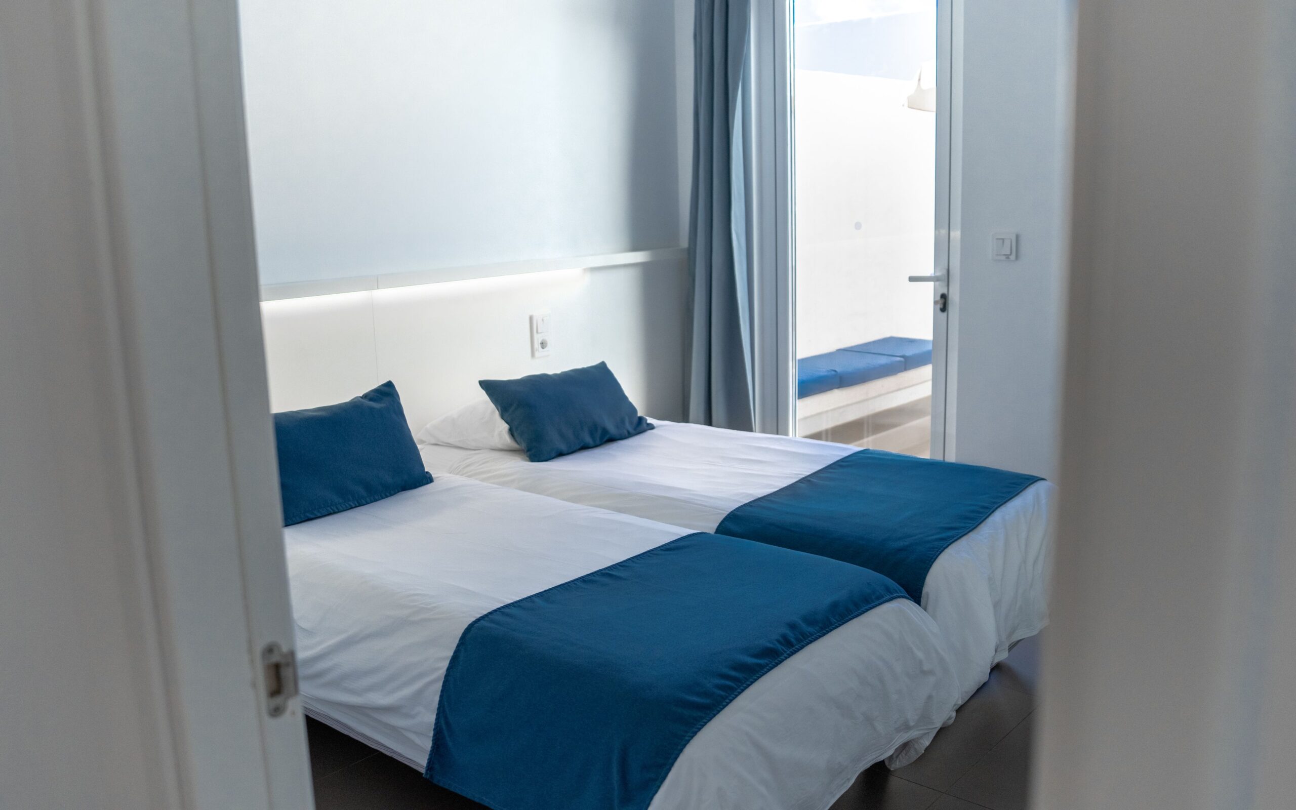 2 single beds with white and blue beddng in a bedroom with white walls and a glass patio door.