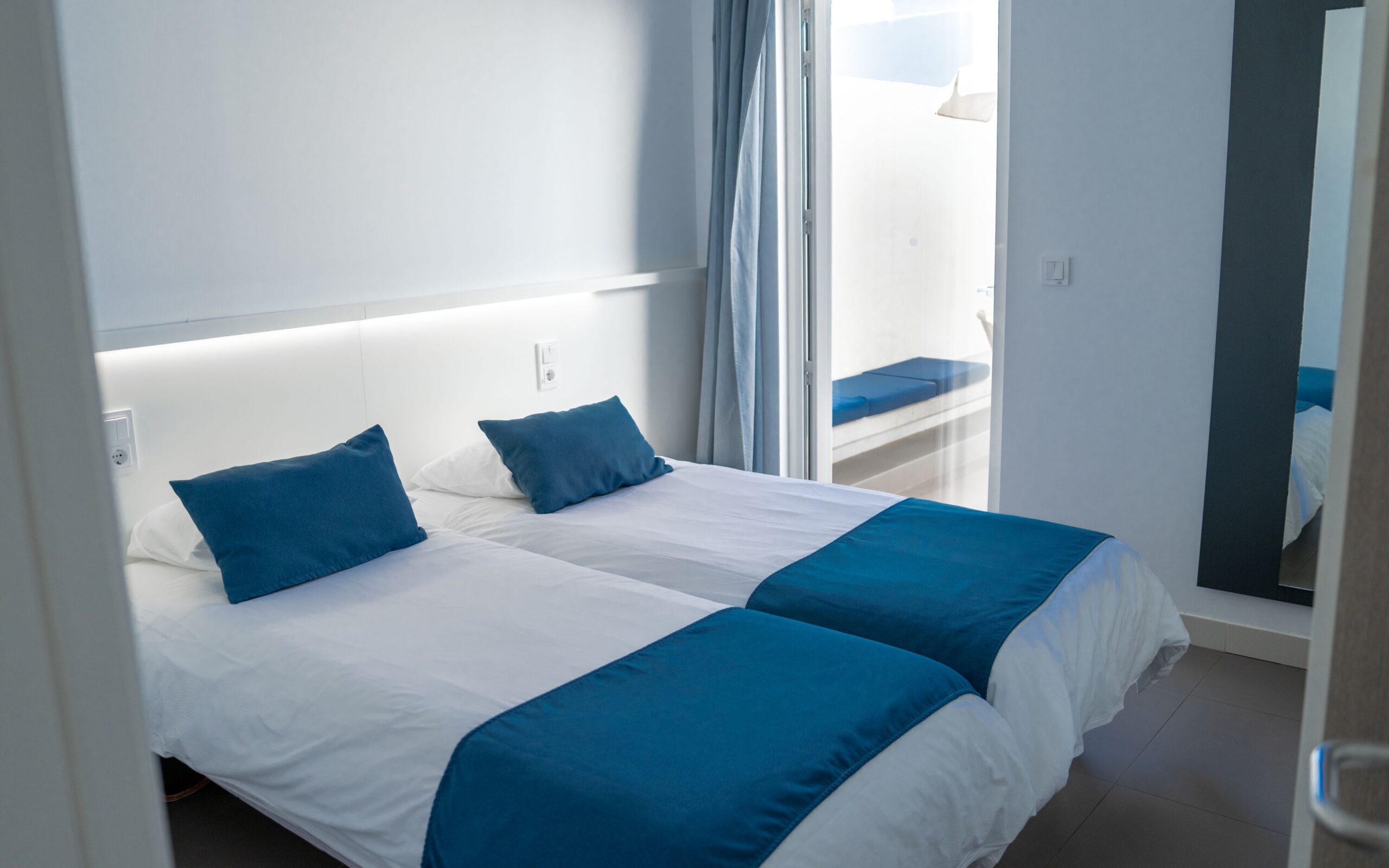 2 single beds with white and blue beddng in a bedroom with white walls and a glass patio door.