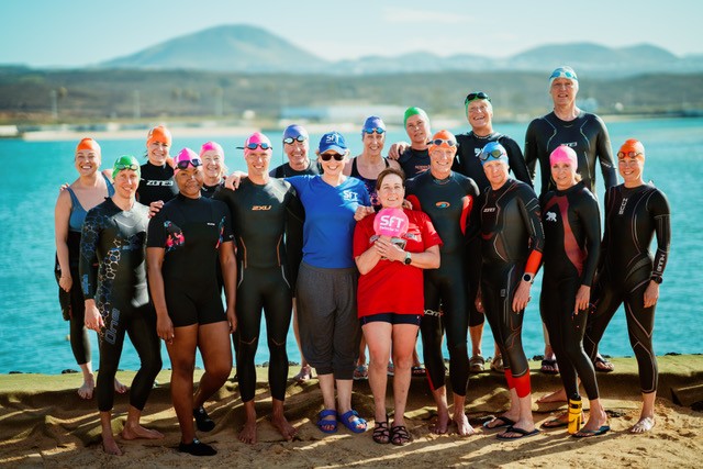 Group of open water swimmers in wetsuits and swim caps standing on the beach at Club La Santa with an instructor in a red SwimForTri top.