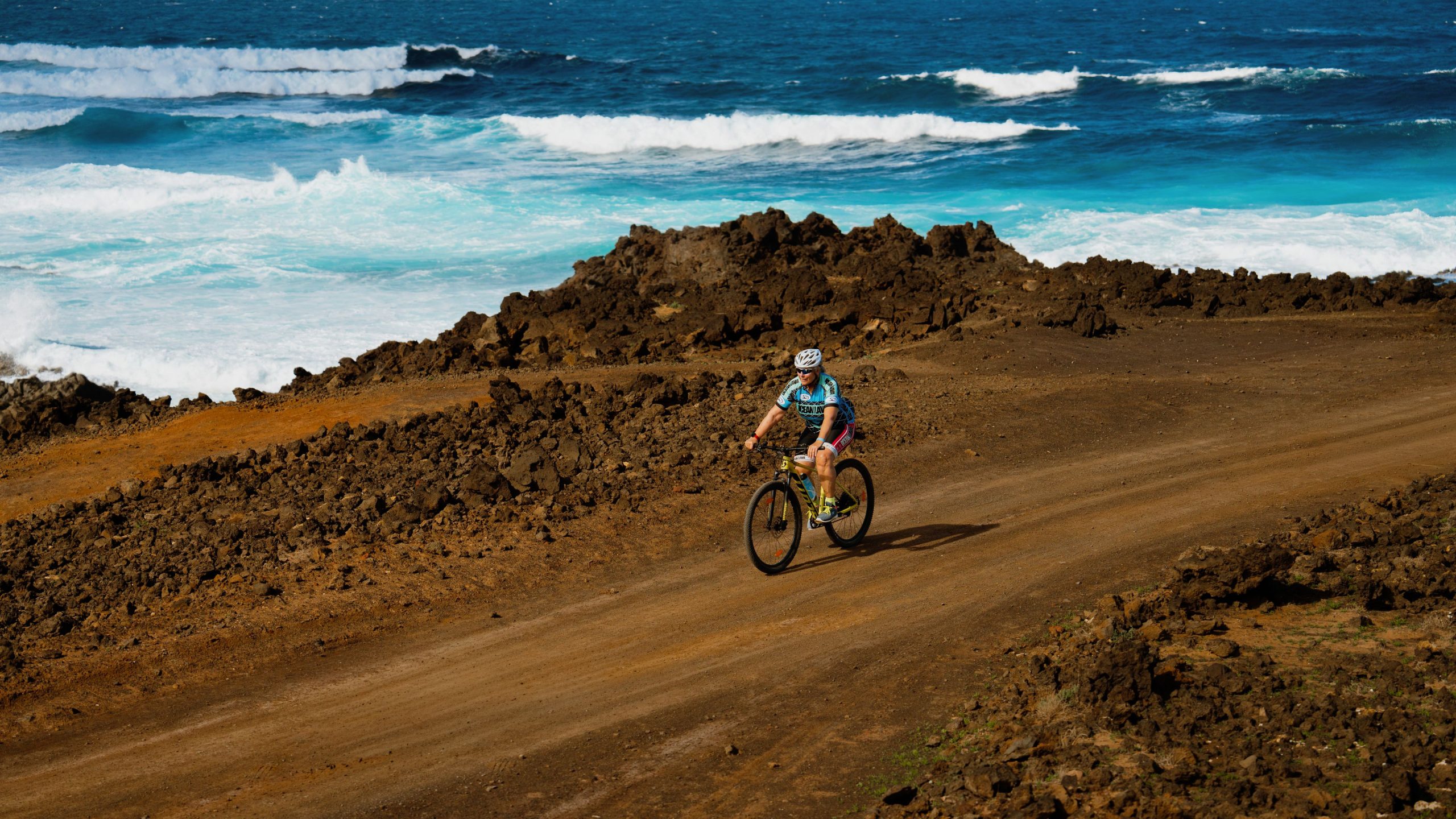 A woman wearing a white bike helmet and blue cycling suit, riding a bike with the sea in the background.