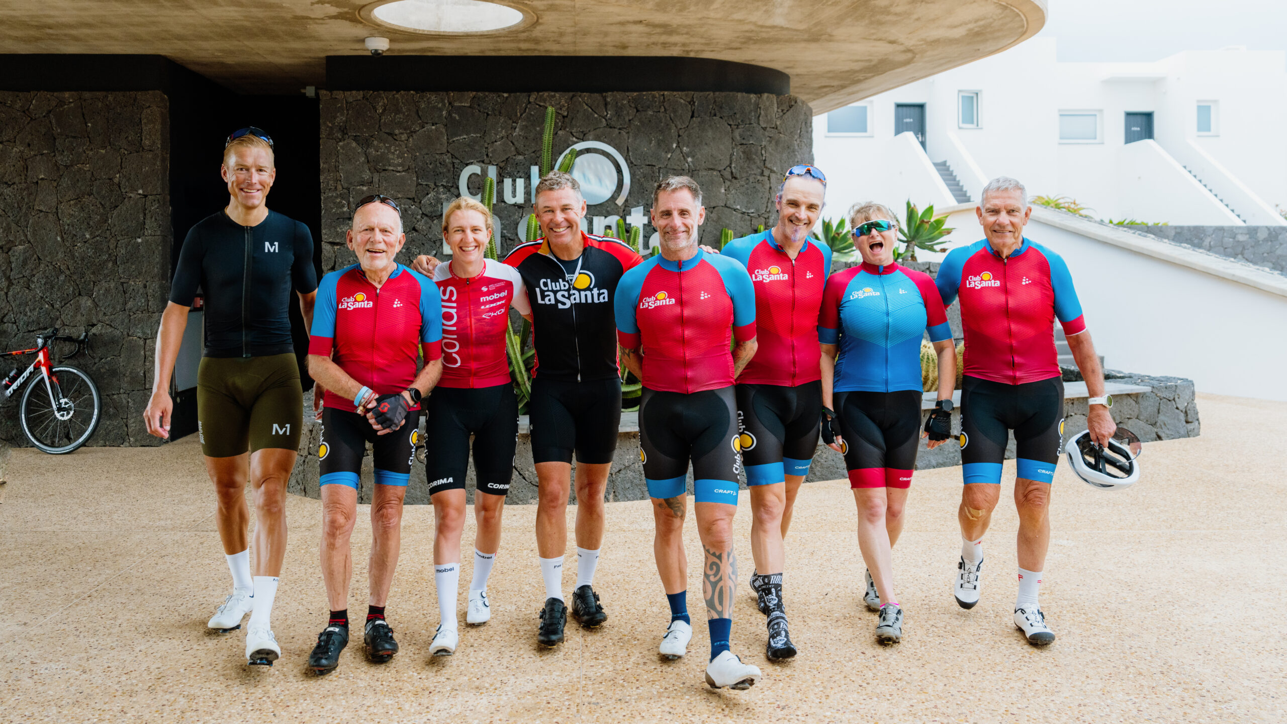 A group of cyclists, mostly wearing red and blue Club La Santa branded cycling suits, posing for a photo with their instructor at International Bike Week.