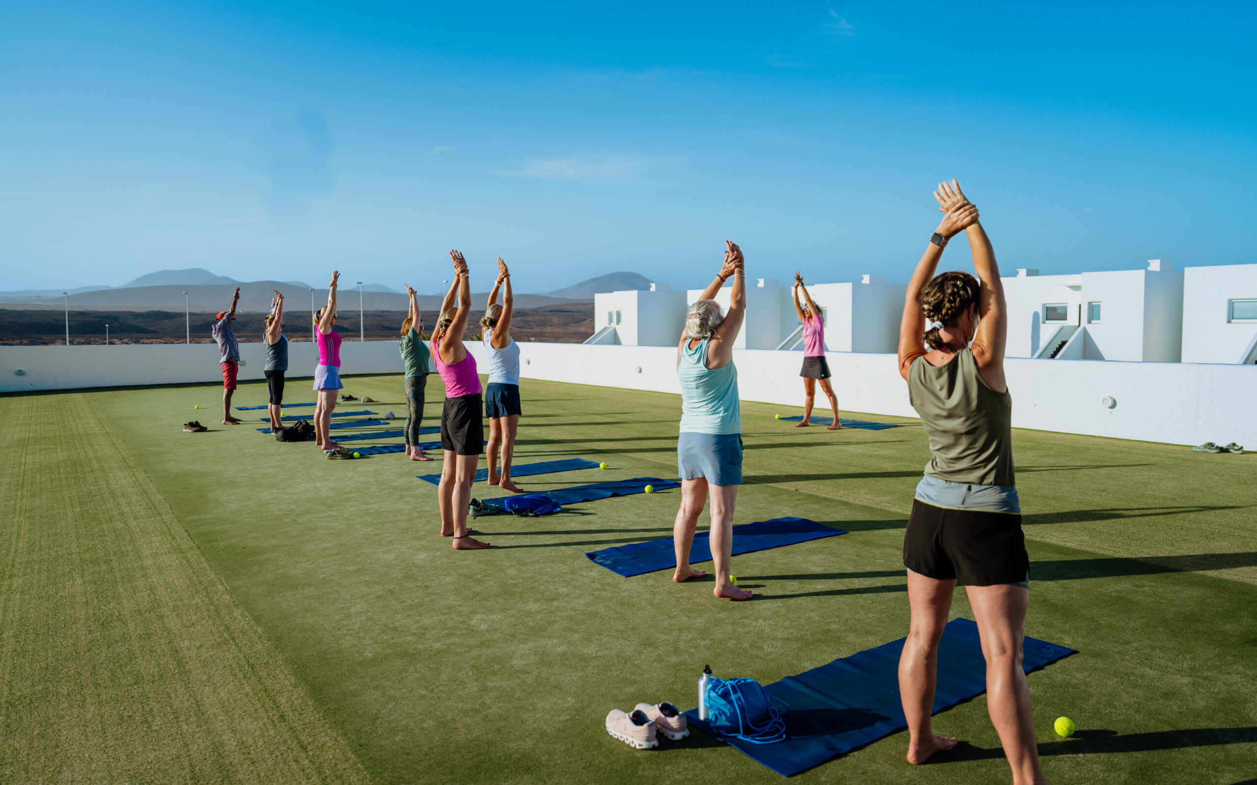 A group of adults stood on the astro-turfed roof at Club La Santa. They are following the pilates instructor and stretching their arms in the air.