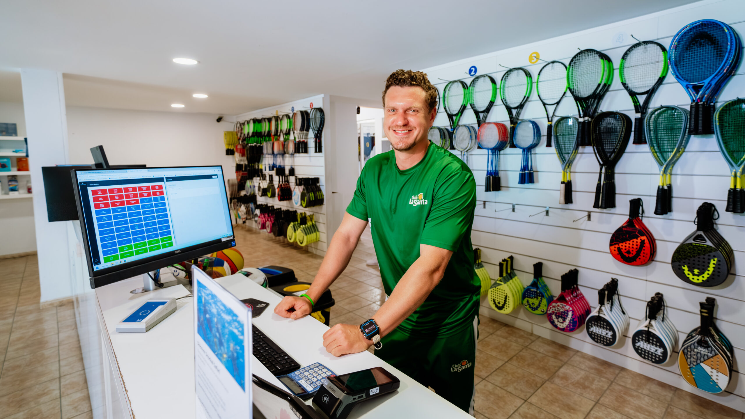 A member of staff at Club La Santa stood behind the Sports Booking desk. There is a variety of padel, tennis and badminton rackets on the wall behind him.