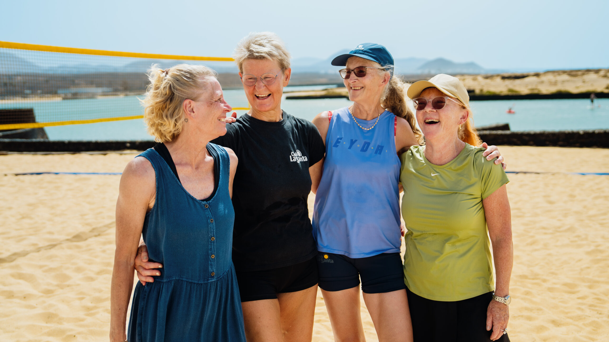 A group of four women with their arms around each other, stood on the beach volleyball court at Club La Santa