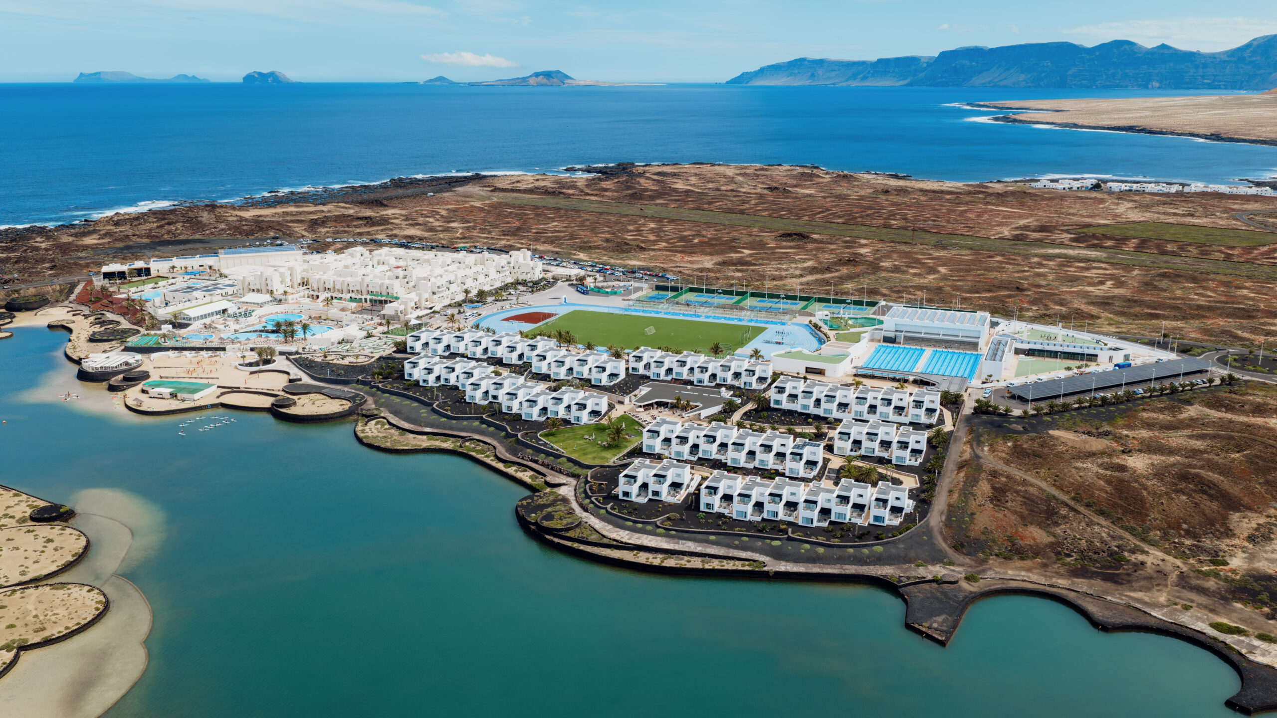 This image shows an aerial view of a coastal resort complex. The resort is situated on a peninsula with the ocean on one side and a large body of water, likely a lagoon, on the other. The complex features numerous white buildings arranged in rows, suggesting residential or hotel accommodations. There are also several swimming pools and what appears to be a sports field with running tracks. The surrounding landscape includes rocky outcrops, green fields, and distant mountains. The clear blue sky and calm waters contribute to the serene and luxurious atmosphere of the resort.