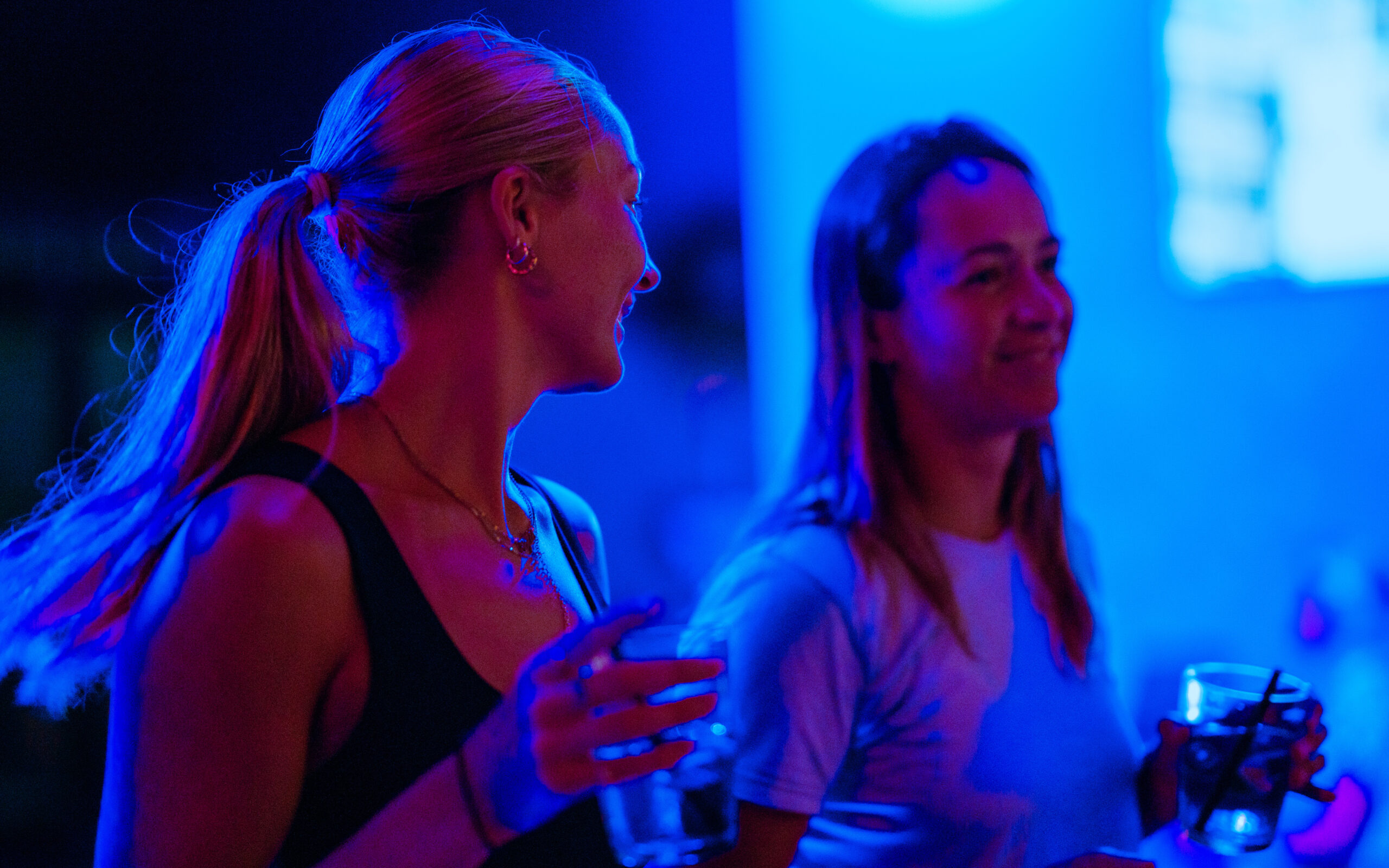 This image shows two individuals standing close to each other in a dimly lit environment with blue lighting. Both individuals are holding glasses with drinks in them. The individual on the left has their hair tied back in a ponytail and is wearing a sleeveless top. The individual on the right has long hair and is wearing a t-shirt. They appear to be engaged in a conversation or enjoying the atmosphere around them.