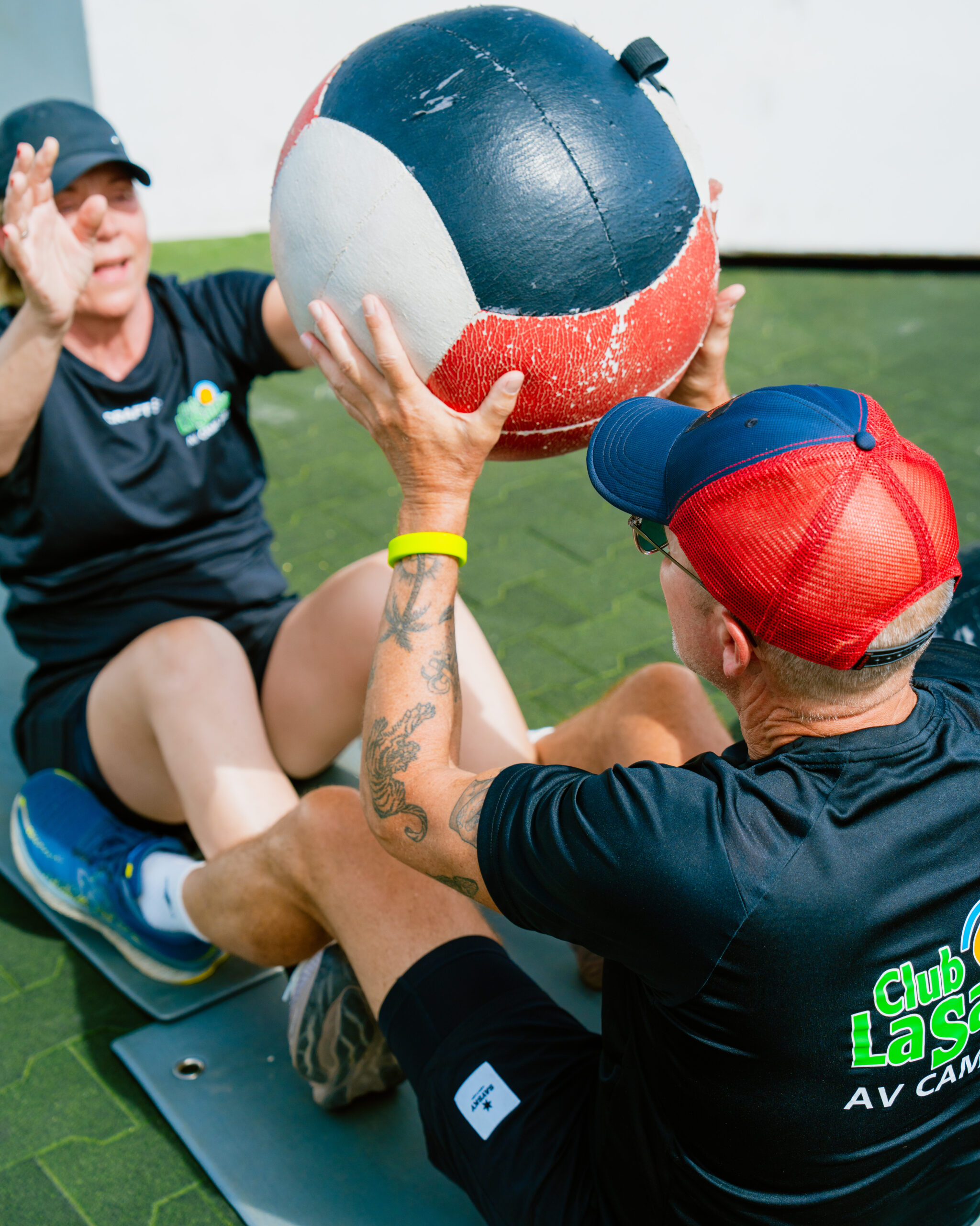 Two people sat on the floor facing each other. They are doing an abdominal exercise whether they do sit-ups, passing a weighted ball between them.