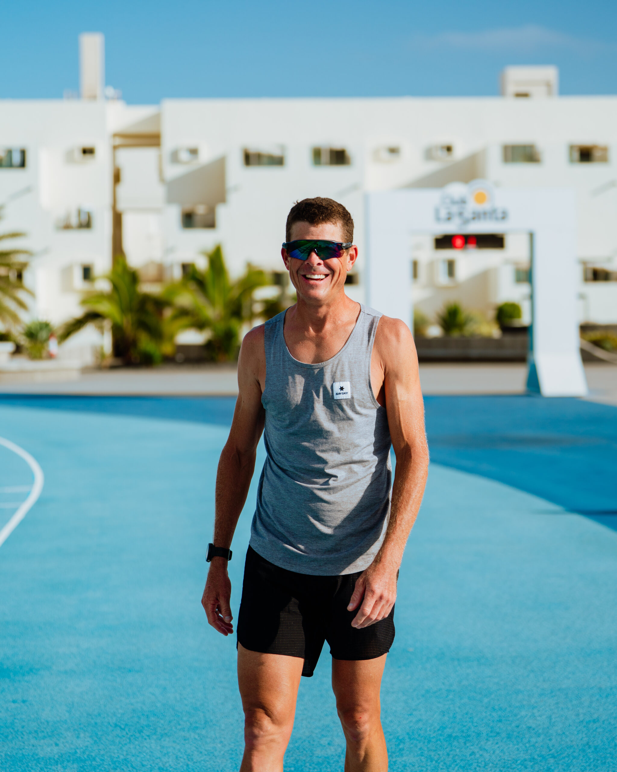 This image shows a person standing on a blue track, smiling and posing for a photo. He is wearing a gray tank top with a small white logo on the left side of his chest, black shorts, and sunglasses.