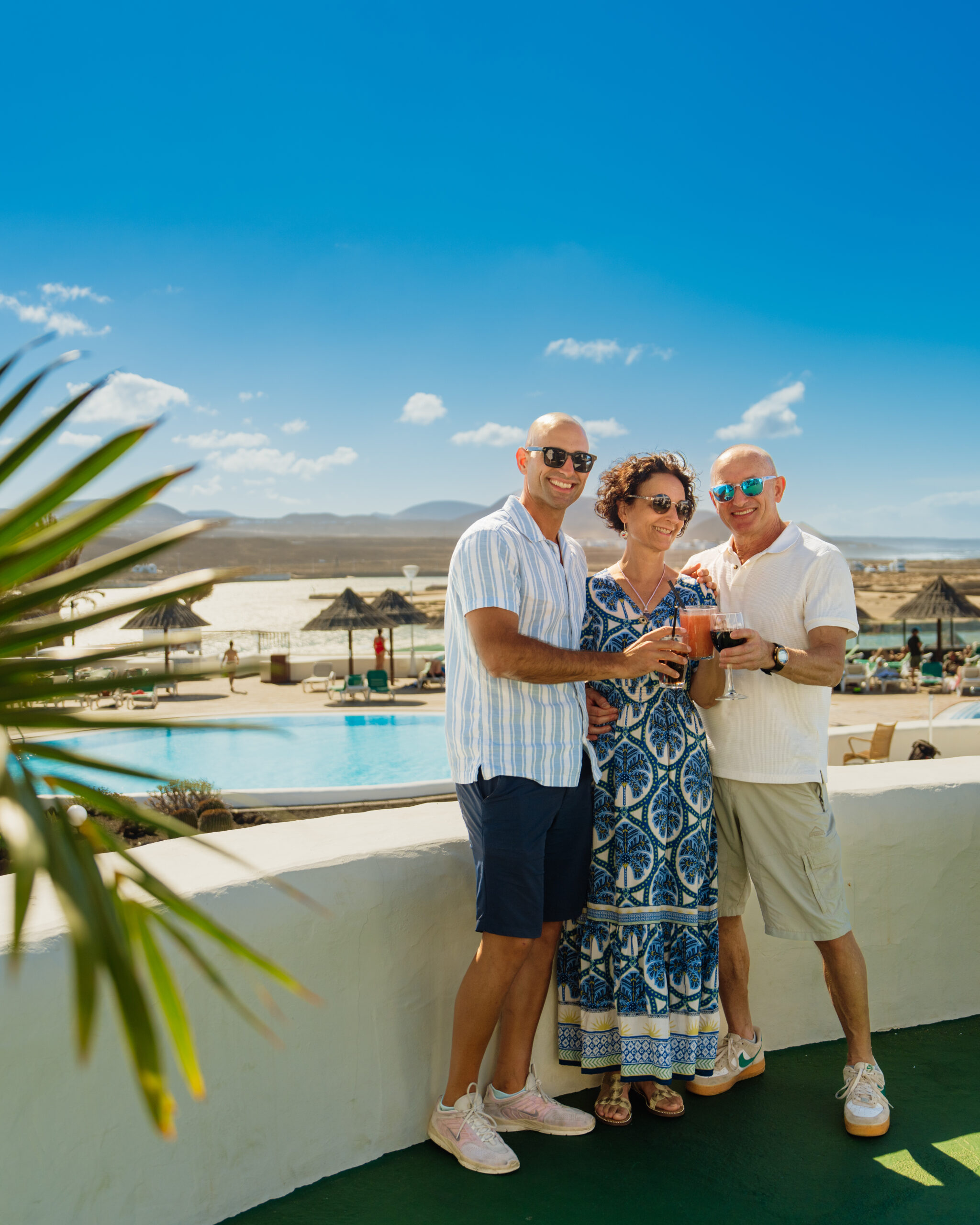 three individuals standing together on a balcony overlooking a resort with a swimming pool. They are all smiling and appear to be posing for a photograph. They are holding glasses with drinks in them. In the background, there are lounge chairs and umbrellas around the pool, and a clear blue sky above.
