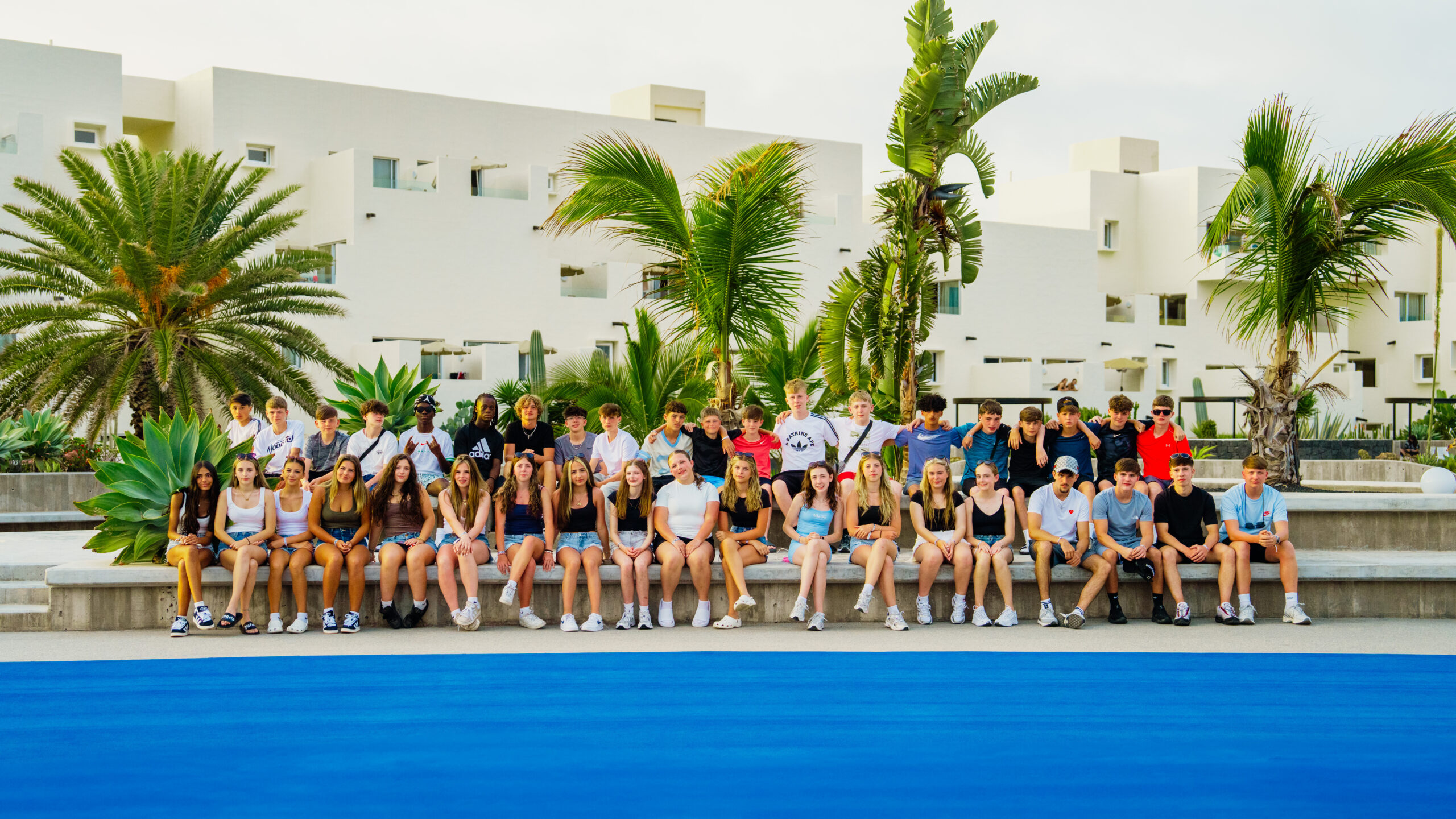 A school group having a group photo at Club La Santa. They are positioned in 2 rows, with the front row sat down on a wall, and the back row stood up behind them. There are palm trees and white villas in the background.