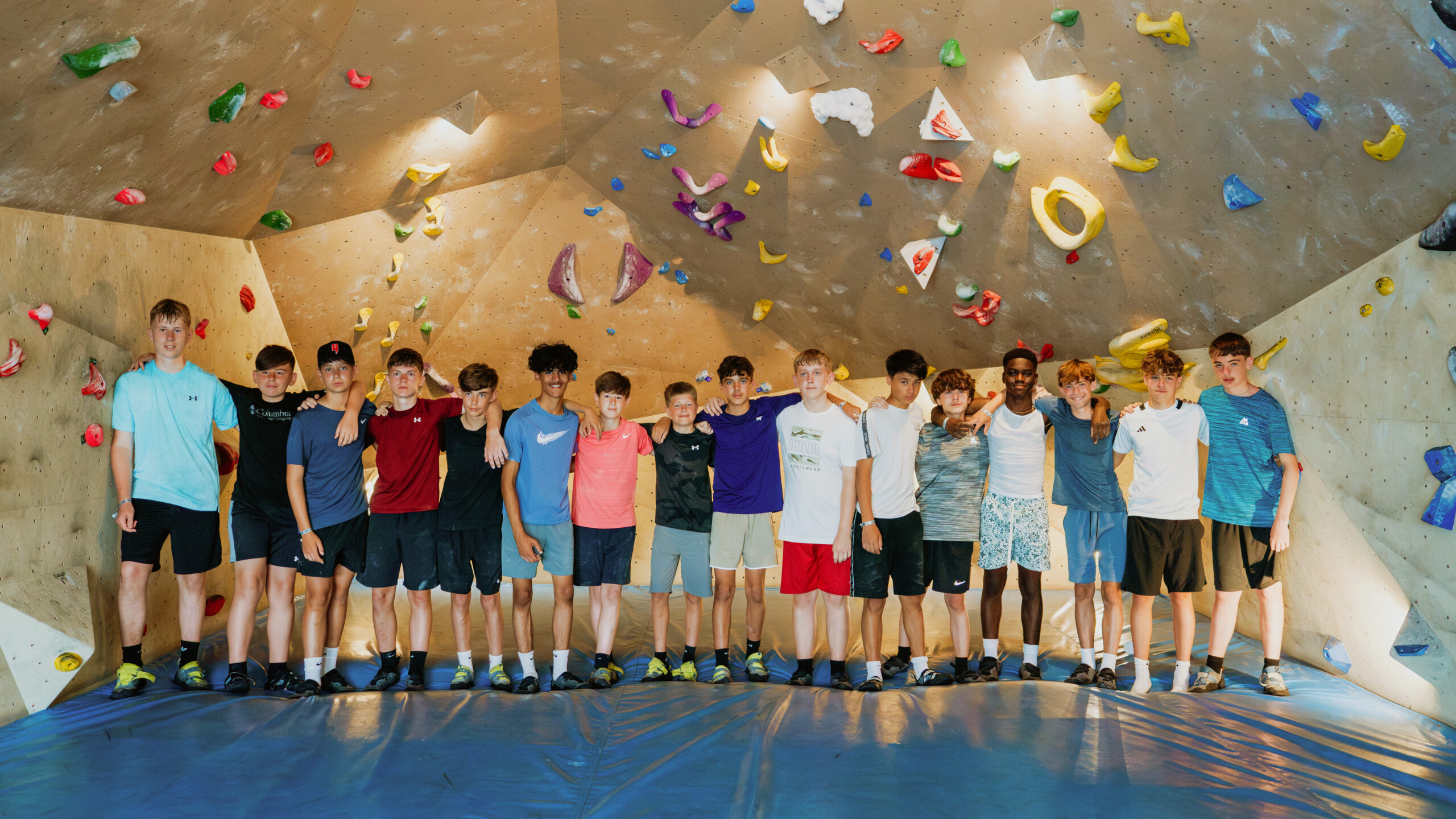 A group of school kids stood posing for a photo with their arms around each other. They are in the Bouldering Centre at Club La Santa.