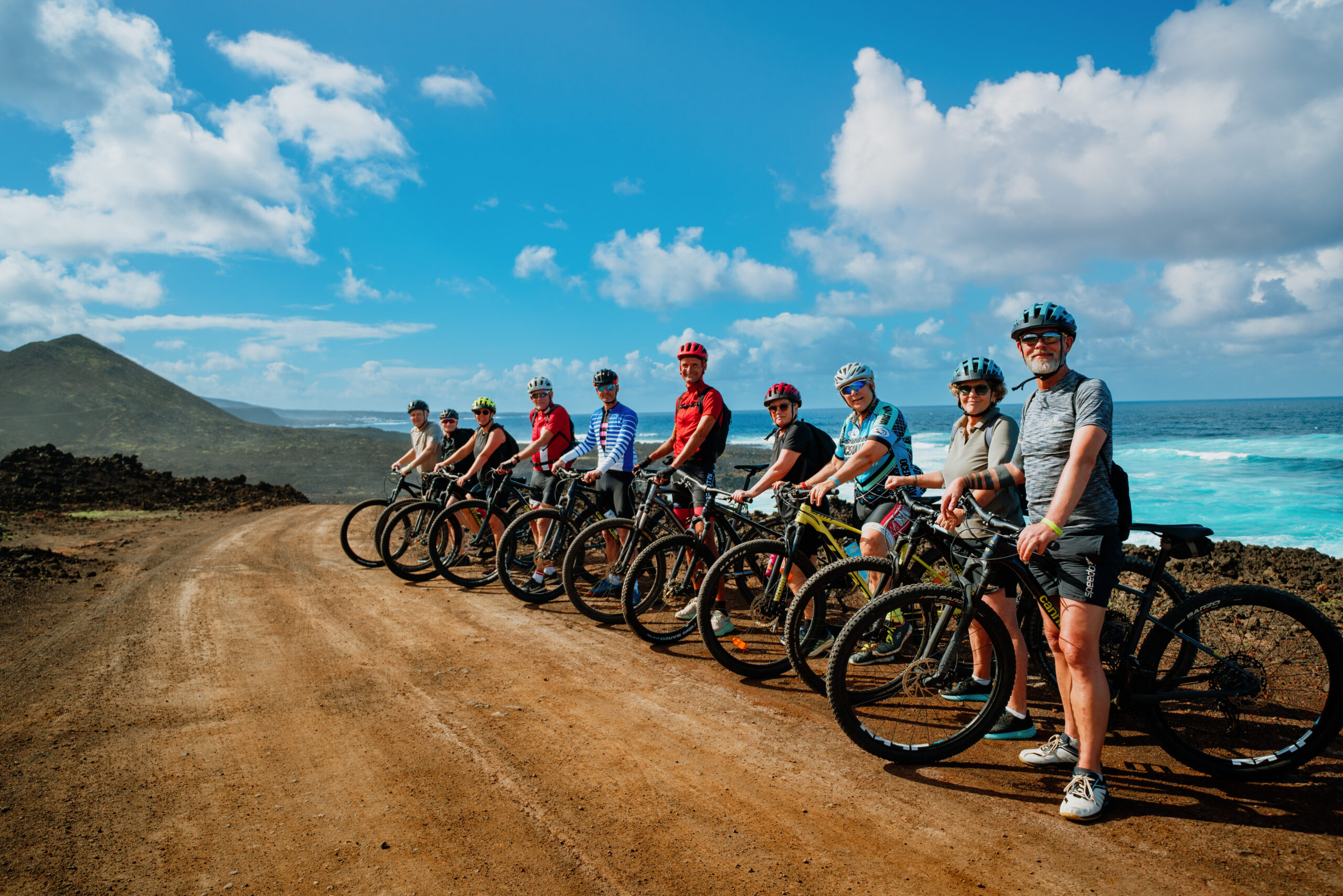 This image shows a group of cyclists lined up on a dirt road near the ocean. They are all wearing helmets and cycling gear, with some individuals sporting sunglasses. The bicycles are arranged in a row, and the participants appear to be preparing for a ride. The ocean is visible in the background, with waves crashing against the shore. The sky is partly cloudy, and there is a mountain visible in the distance. The scene conveys a sense of adventure and outdoor activity.
