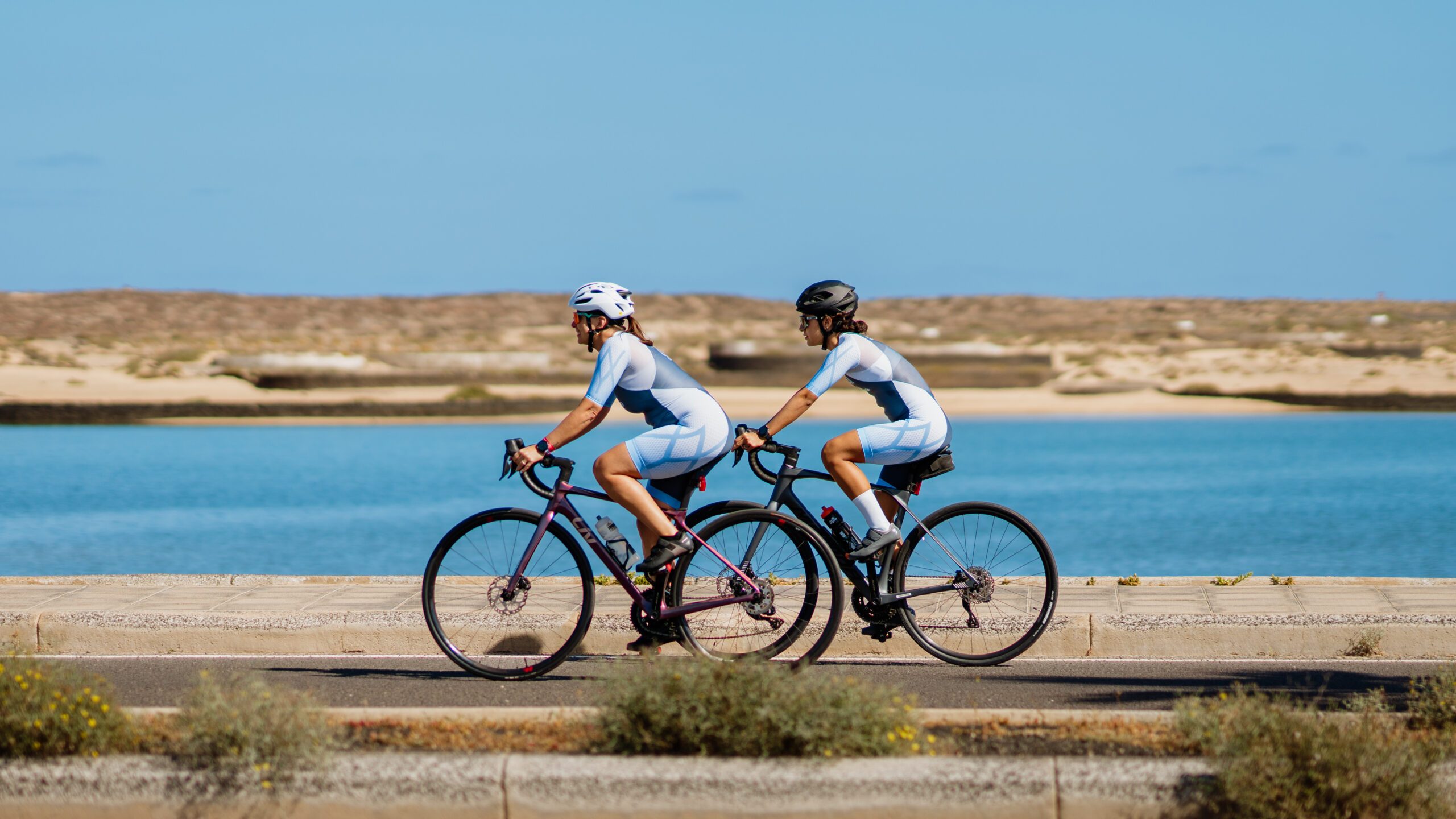 This image shows two individuals riding bicycles side by side on a road near a body of water under a clear blue sky. Both cyclists are wearing helmets and cycling suits, with one cyclist in a white helmet and the other in a black helmet. The bicycles are modern road bikes with water bottles attached to the frames. The road is bordered by a concrete sidewalk on one side and a grassy area with some low vegetation on the other. In the background, there is a large expanse of water and a sandy, possibly coastal, landscape.